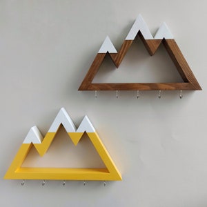 May include: Two wooden mountain-shaped key racks with hooks. The top rack is brown with white peaks and the bottom rack is yellow with white peaks.