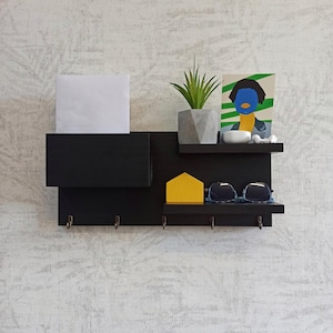 Puede incluir: Estantería de pared negra con ganchos, una pequeña estantería y una ranura para el correo. La estantería tiene un objeto amarillo en forma de casa, gafas de sol y un objeto rectangular blanco. Una planta en maceta y una impresión enmarcada están en la estantería superior.