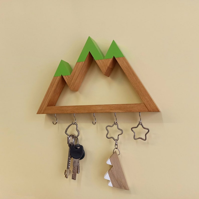 May include: Wooden key holder shaped like a mountain range with green peaks. It has five hooks and is decorated with a star-shaped key chain and a small wooden tree.