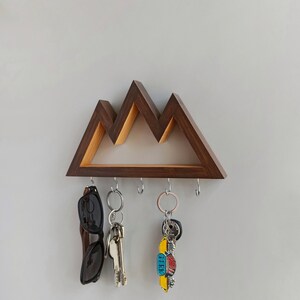 Puede incluir: Porta llaves de madera con forma de montaña con cinco ganchos. Gafas de sol y llaves están colgadas de los ganchos.