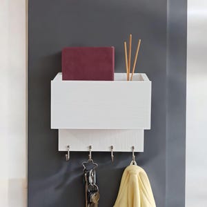 May include: White wooden wall mounted key holder with a shelf and hooks. The shelf has a burgundy book on it and a set of wooden sticks in a glass vase. There are five hooks on the bottom.