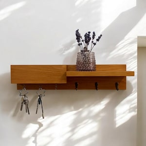 May include: A wooden wall-mounted shelf with a key rack and a small shelf on top. The shelf is light brown and has five hooks for keys. There is a vase with dried lavender on the top shelf.