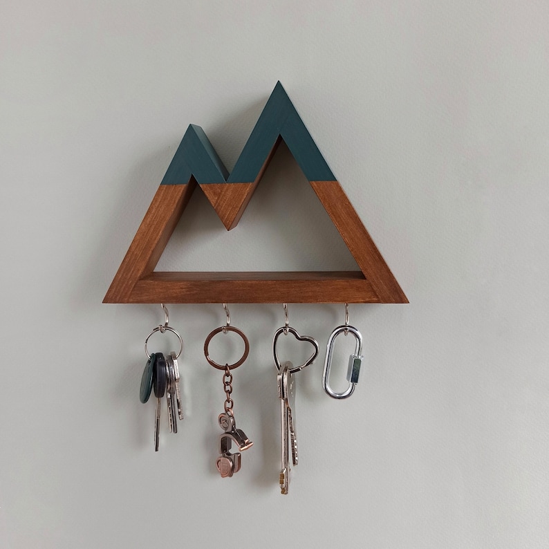May include: Wooden key holder shaped like a mountain range with a dark teal top and a light brown base. Five hooks are attached to the bottom of the holder, and there are keys and a carabiner hanging from the hooks.