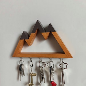 May include: Wooden key holder shaped like a mountain range with a brown wood base and a dark gray top. Five keys are hanging from hooks on the bottom of the holder.