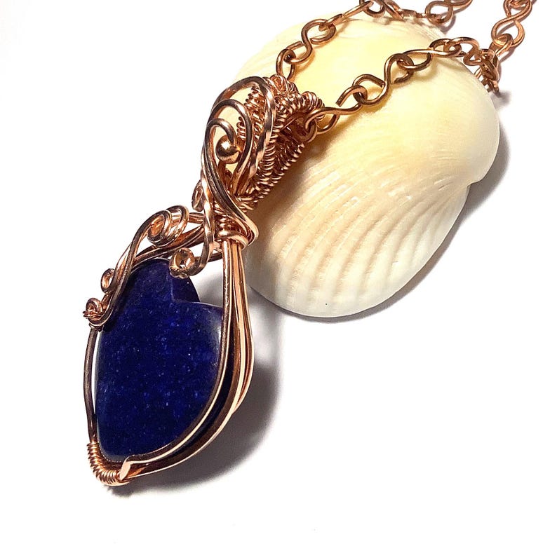 Puede incluir: Un collar con colgante artesanal de alambre de cobre con una piedra preciosa azul oscuro. El colgante tiene un intrincado dise&ntilde;o envuelto en alambre, con forma de l&aacute;grima. La cadena del collar tambi&eacute;n es de color cobre. El colgante est&aacute; sobre una concha blanca.