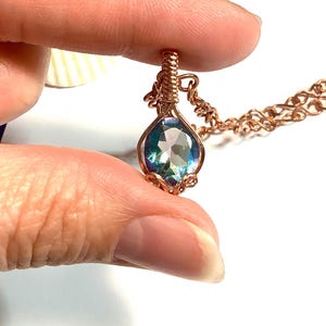 Puede incluir: Un colgante en forma de l&aacute;grima con una piedra preciosa facetada de tonos azules y morados iridiscentes, engastada en alambre de cobre enrollado. El colgante est&aacute; unido a una cadena de cobre a juego, con el alambre formando una argolla.