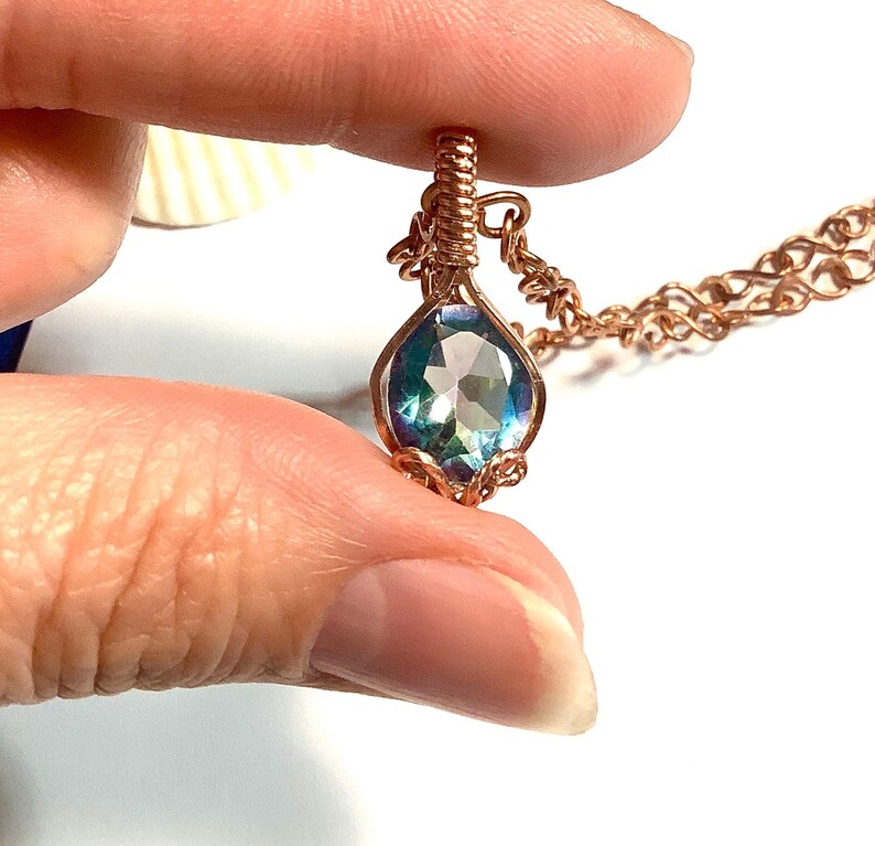 Puede incluir: Un colgante en forma de l&aacute;grima con una piedra preciosa facetada de tonos azules y morados iridiscentes, engastada en alambre de cobre enrollado. El colgante est&aacute; unido a una cadena de cobre a juego, con el alambre formando una argolla.