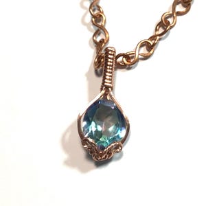 Puede incluir: Un collar con un colgante de piedra preciosa iridiscente en tonos azul y violeta con forma de l&aacute;grima, engastado en alambre de cobre. El colgante cuelga de una cadena de cobre compuesta por eslabones entrelazados en forma de infinito.