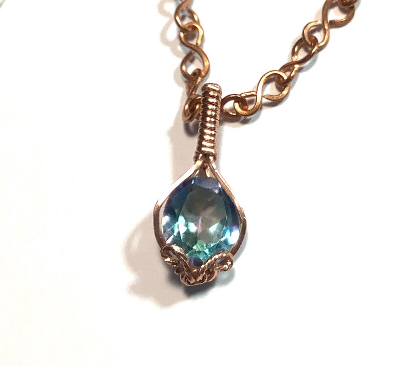 Puede incluir: Un collar con un colgante de piedra preciosa iridiscente en tonos azul y violeta con forma de l&aacute;grima, engastado en alambre de cobre. El colgante cuelga de una cadena de cobre compuesta por eslabones entrelazados en forma de infinito.