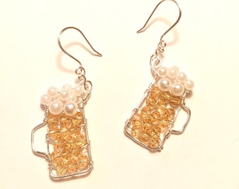 May include: A pair of silver wire earrings shaped like beer steins. The steins are filled with amber-colored beads and topped with white pearl beads.
