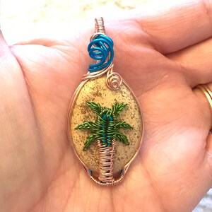May include: A copper wire wrapped pendant featuring a palm tree design on a beige stone. The tree is made of green and blue wire. The pendant has a blue wire loop at the top.