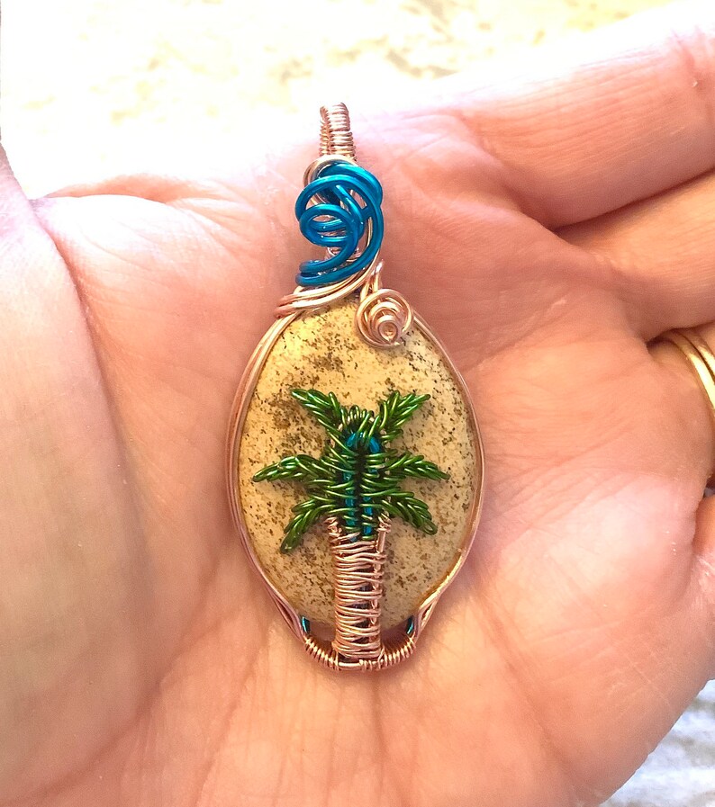 May include: A copper wire wrapped pendant featuring a palm tree design on a beige stone. The tree is made of green and blue wire. The pendant has a blue wire loop at the top.