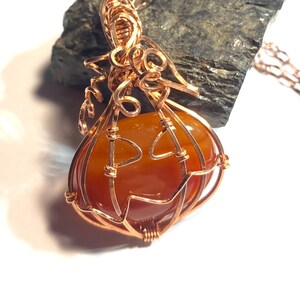 May include: A wire-wrapped pendant featuring a smooth, rounded orange carnelian stone encased in copper wire shaped to resemble a pumpkin. The copper wire forms a stem with curly tendrils at the top and a jack-o'-lantern face on the front surface.
