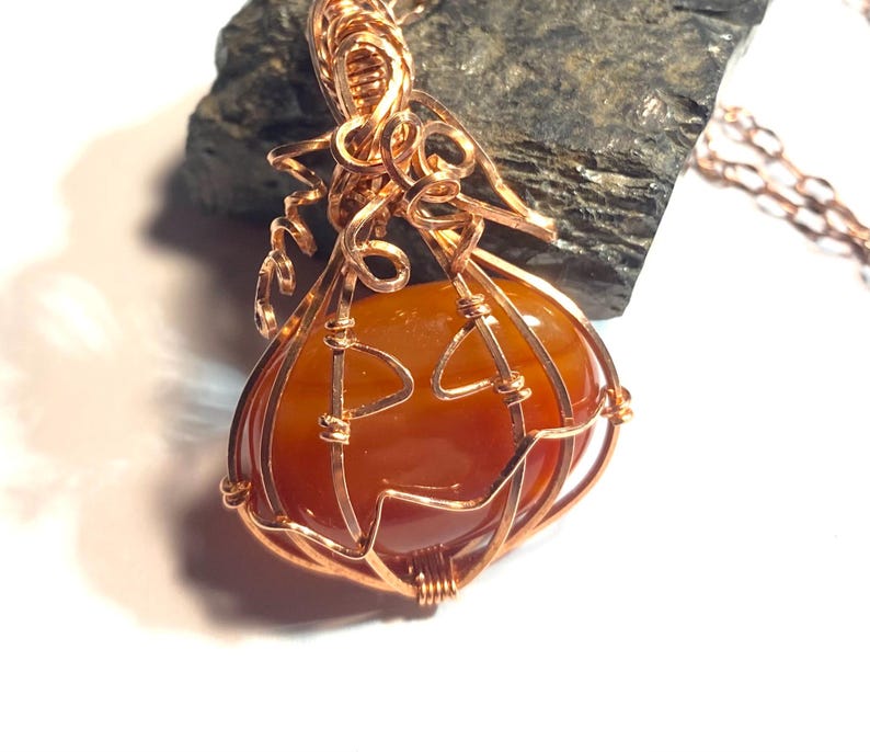 May include: A wire-wrapped pendant featuring a smooth, rounded orange carnelian stone encased in copper wire shaped to resemble a pumpkin. The copper wire forms a stem with curly tendrils at the top and a jack-o'-lantern face on the front surface.