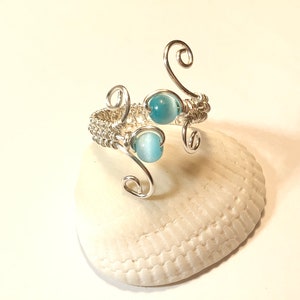 May include: A silver wire wrapped ring with two light blue gemstone beads. The ring is on a white seashell.