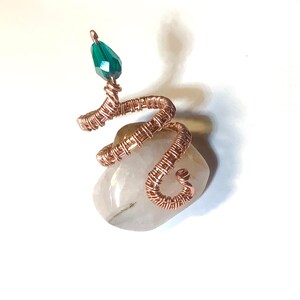 以下が含まれることがあります： 滑らかな明るい色の石に巻き付いた、ヘビの形をした銅線リング。ヘビの頭には、ファセット加工されたエメラルドグリーンのビーズが付いています。リングのデザインは複雑で、ワイヤーはヘビの体を形成するように慎重に巻かれています。