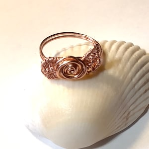 May include: A rose-gold wire-wrapped ring with a single rose design. The ring is sitting on a white seashell.