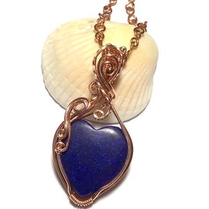 Puede incluir: Un collar con colgante de alambre de color cobre con una piedra en forma de coraz&oacute;n azul oscuro. El colgante cuelga de una delicada cadena. El collar se muestra sobre un fondo blanco.