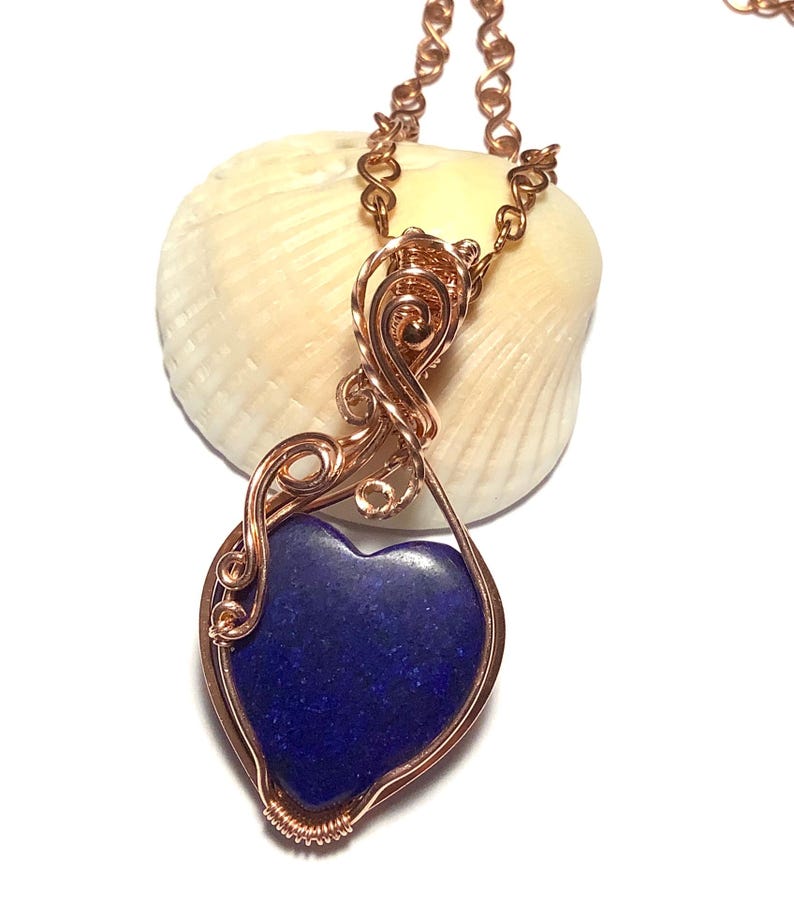 Puede incluir: Un collar con colgante de alambre de color cobre con una piedra en forma de coraz&oacute;n azul oscuro. El colgante cuelga de una delicada cadena. El collar se muestra sobre un fondo blanco.