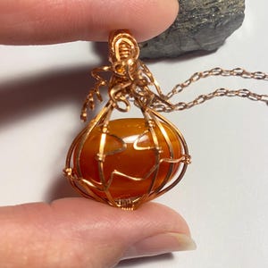 May include: A wire-wrapped pendant featuring a smooth, rounded orange carnelian stone encased in copper wire shaped like a pumpkin. The design includes curled wire tendrils at the top and a matching copper chain attached to the bail.