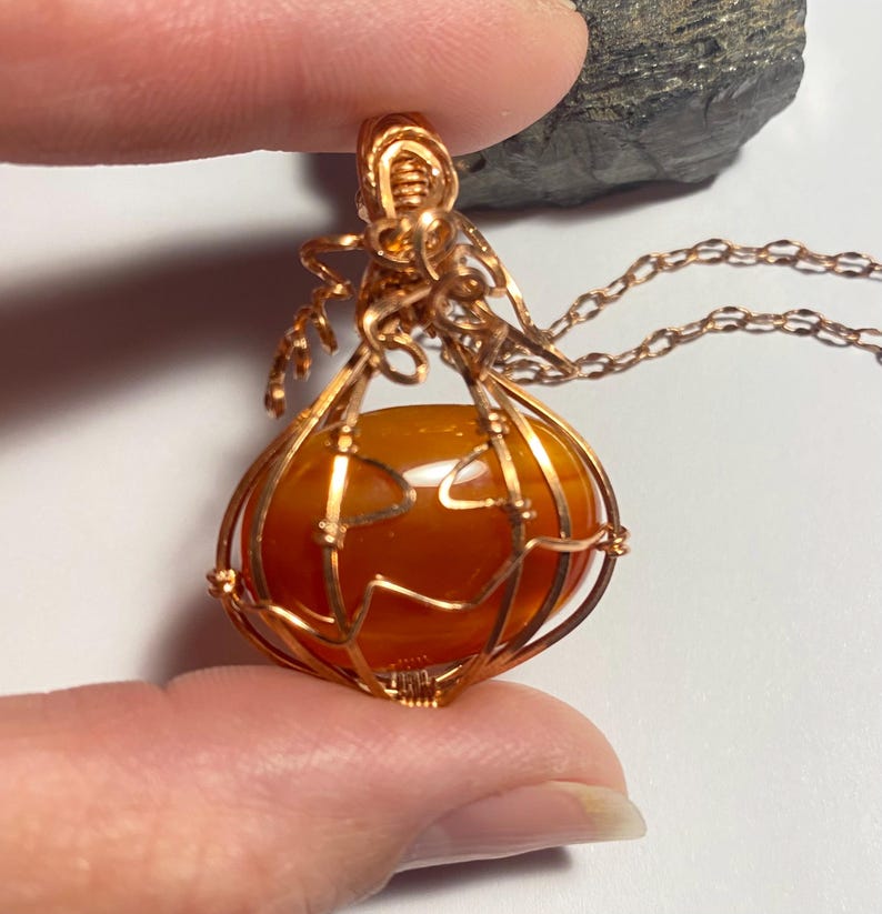 May include: A wire-wrapped pendant featuring a smooth, rounded orange carnelian stone encased in copper wire shaped like a pumpkin. The design includes curled wire tendrils at the top and a matching copper chain attached to the bail.