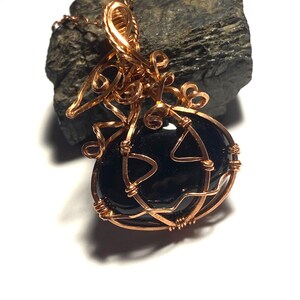 May include: A pendant featuring a smooth, polished black oval stone encased in intricate copper wire wrapping. The wire forms decorative loops and swirls around the stone, extending upward into a bail for a chain, with a metallic copper finish.