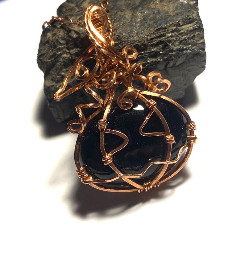 May include: A pendant featuring a smooth, polished black oval stone encased in intricate copper wire wrapping. The wire forms decorative loops and swirls around the stone, extending upward into a bail for a chain, with a metallic copper finish.