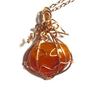 May include: A wire-wrapped pendant necklace featuring a smooth, round, orange carnelian stone encased in copper wire shaped like a pumpkin. The wire forms a jack-o'-lantern face on the front of the stone, with coiled wire tendrils at the top.