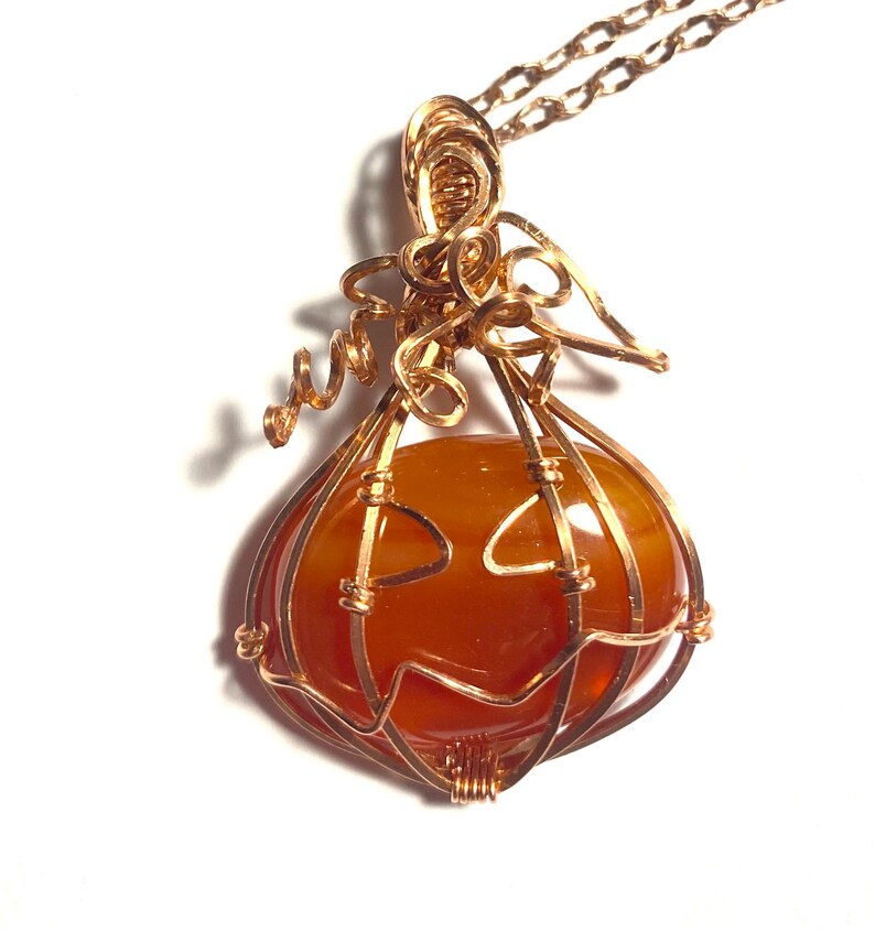 May include: A wire-wrapped pendant necklace featuring a smooth, round, orange carnelian stone encased in copper wire shaped like a pumpkin. The wire forms a jack-o'-lantern face on the front of the stone, with coiled wire tendrils at the top.