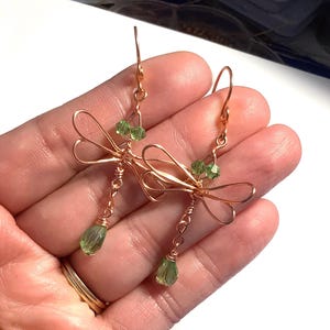May include: Copper wire dragonfly earrings with green crystal accents. The earrings feature a delicate dragonfly design with wire wings and a teardrop-shaped green crystal pendant. The earrings are held up by a copper hook.