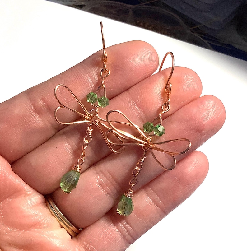 May include: Copper wire dragonfly earrings with green crystal accents. The earrings feature a delicate dragonfly design with wire wings and a teardrop-shaped green crystal pendant. The earrings are held up by a copper hook.