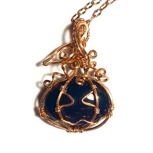 May include: A wire-wrapped pendant necklace featuring a dark, smooth, rounded stone encased in copper-colored wire shaped like a pumpkin with a stem and curly tendrils. The pendant hangs from a matching copper-colored metal chain link necklace.