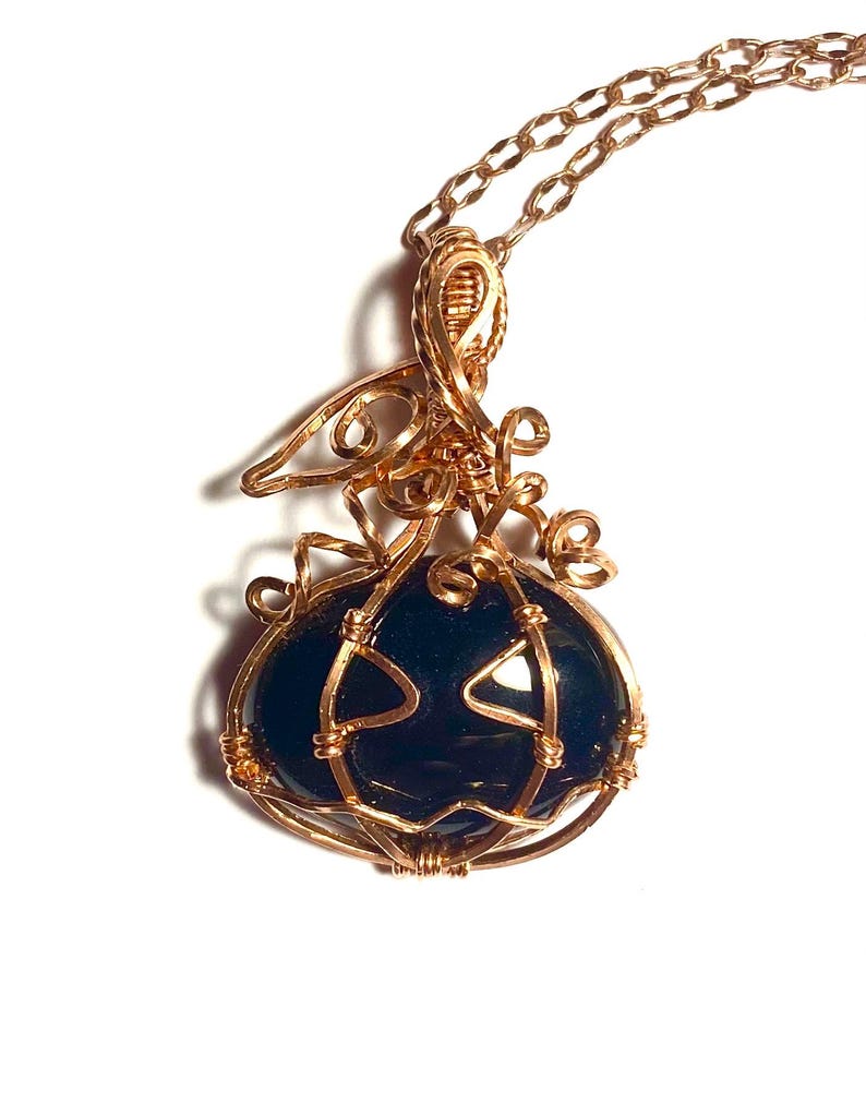 May include: A wire-wrapped pendant necklace featuring a dark, smooth, rounded stone encased in copper-colored wire shaped like a pumpkin with a stem and curly tendrils. The pendant hangs from a matching copper-colored metal chain link necklace.