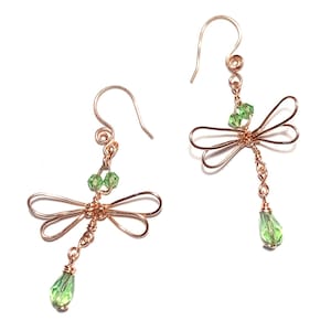 May include: A pair of copper-colored wire earrings, each shaped like a dragonfly. The dragonfly's wings are formed from looped wire, with green crystal beads at the body and a teardrop-shaped green crystal dangling below.