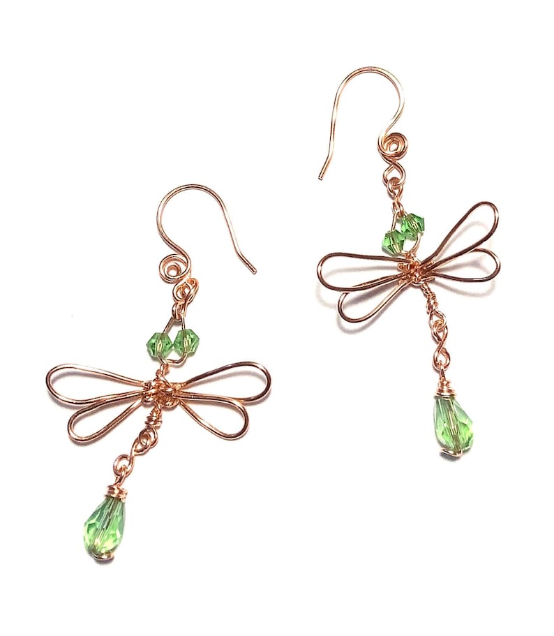 May include: A pair of copper-colored wire earrings, each shaped like a dragonfly. The dragonfly's wings are formed from looped wire, with green crystal beads at the body and a teardrop-shaped green crystal dangling below.