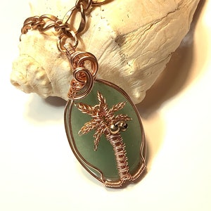 May include: A copper wire wrapped pendant featuring a green stone with a copper wire palm tree design. The palm tree has two small gold beads for coconuts.