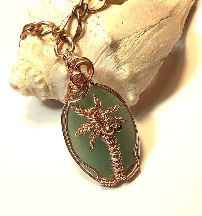 May include: A copper wire wrapped pendant featuring a green stone with a copper wire palm tree design. The palm tree has two small gold beads for coconuts.