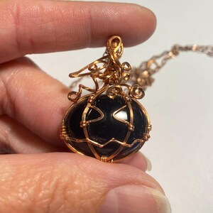 May include: A pendant necklace featuring a smooth, black, oval-shaped stone encased in copper wire. The wire is intricately wrapped to form the shape of a pumpkin with a stem and curly vines at the top, suspended from a matching copper chain.