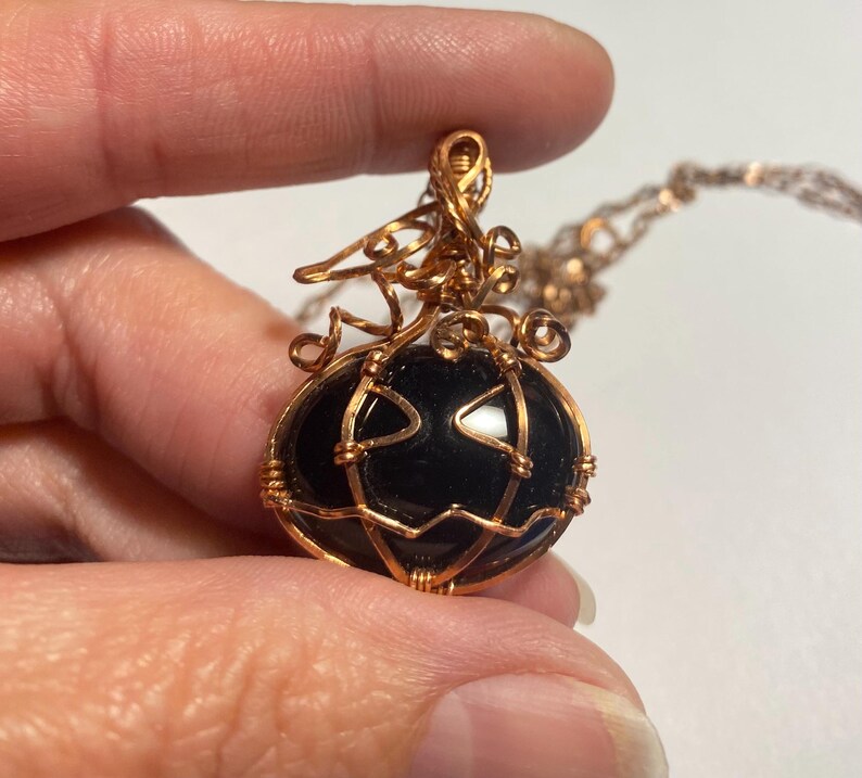 May include: A pendant necklace featuring a smooth, black, oval-shaped stone encased in copper wire. The wire is intricately wrapped to form the shape of a pumpkin with a stem and curly vines at the top, suspended from a matching copper chain.