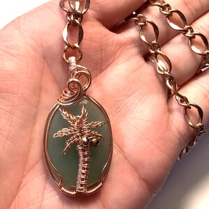 May include: A green stone pendant with a copper wire wrapped palm tree design hangs from a gold chain. The pendant is oval shaped and the palm tree has two coconuts.
