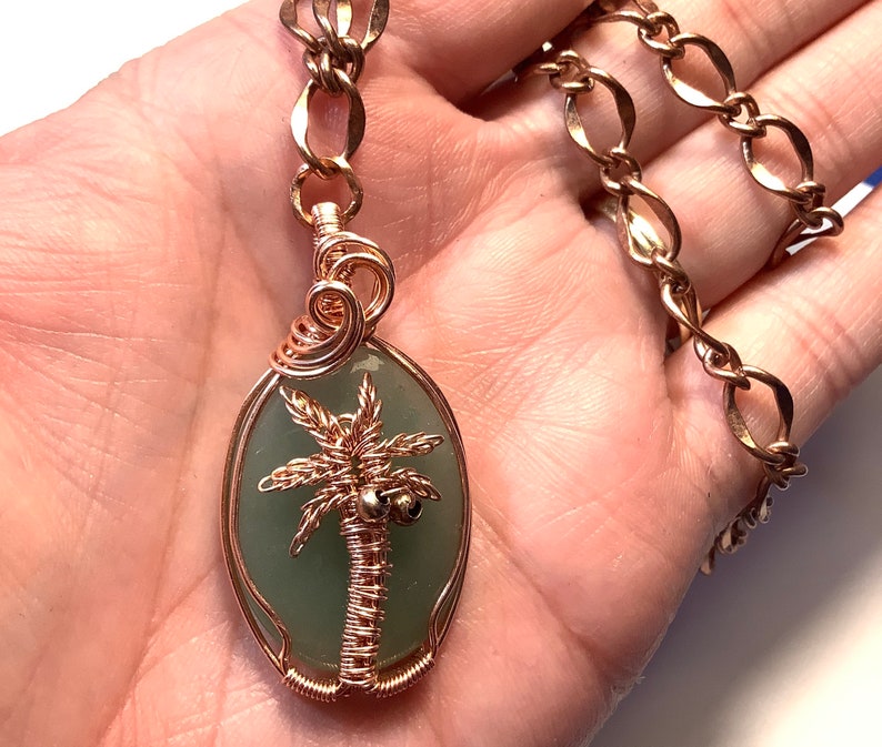 May include: A green stone pendant with a copper wire wrapped palm tree design hangs from a gold chain. The pendant is oval shaped and the palm tree has two coconuts.