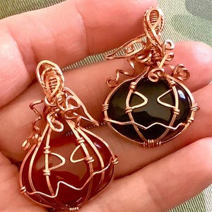 May include: Two wire-wrapped pendants shaped like pumpkins. One pendant features a smooth, dark red stone, and the other features a smooth, black stone. Both are encased in copper-colored wire, forming pumpkin-like ridges and carved facial features.