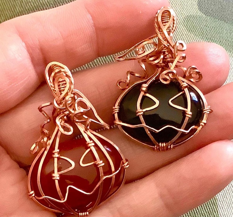 May include: Two wire-wrapped pendants shaped like pumpkins. One pendant features a smooth, dark red stone, and the other features a smooth, black stone. Both are encased in copper-colored wire, forming pumpkin-like ridges and carved facial features.