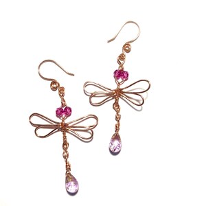 May include: A pair of handcrafted dragonfly earrings. The earrings feature copper-colored wire forming dragonfly shapes, with pink crystal beads and teardrop-shaped pendants. The earrings have a hook closure and are set against a white background.