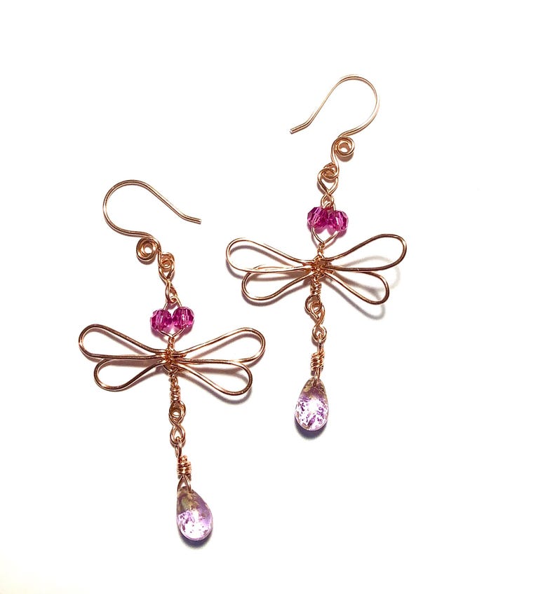 May include: A pair of handcrafted dragonfly earrings. The earrings feature copper-colored wire forming dragonfly shapes, with pink crystal beads and teardrop-shaped pendants. The earrings have a hook closure and are set against a white background.