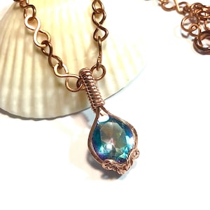 Puede incluir: Un collar con una piedra preciosa facetada en forma de l&aacute;grima de color azul y morado iridiscente, asegurada en un engaste de alambre de cobre enrollado. El colgante cuelga de una cadena de cobre compuesta por eslabones entrelazados en forma de infinito.