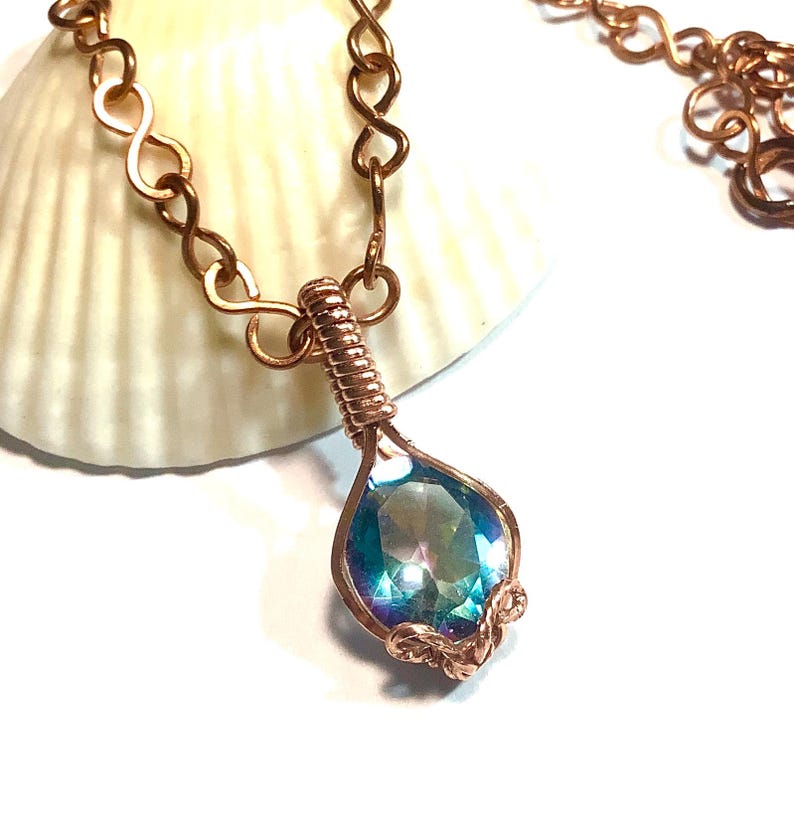 Puede incluir: Un collar con una piedra preciosa facetada en forma de l&aacute;grima de color azul y morado iridiscente, asegurada en un engaste de alambre de cobre enrollado. El colgante cuelga de una cadena de cobre compuesta por eslabones entrelazados en forma de infinito.