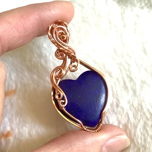 Puede incluir: Un colgante de alambre color cobre con una piedra azul oscuro en forma de coraz&oacute;n. El alambre est&aacute; intrincadamente moldeado con espirales y bucles decorativos, creando un dise&ntilde;o elegante y artesanal. El colgante se sostiene entre los dedos.