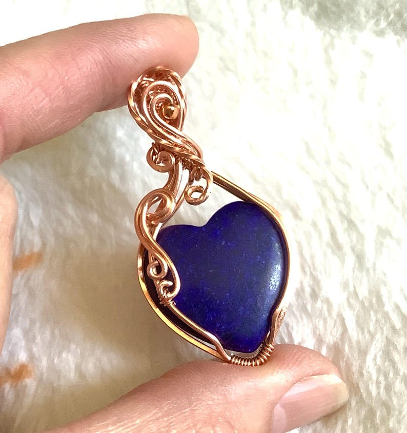Puede incluir: Un colgante de alambre color cobre con una piedra azul oscuro en forma de coraz&oacute;n. El alambre est&aacute; intrincadamente moldeado con espirales y bucles decorativos, creando un dise&ntilde;o elegante y artesanal. El colgante se sostiene entre los dedos.