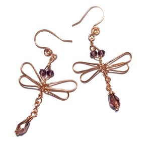 May include: A pair of copper-colored dragonfly earrings. Each earring features a dragonfly design with wire wings, small purple beads, and a dangling faceted purple crystal. The earrings have a hook closure.